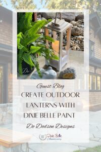 A white and brown wooden lantern wit a yellow lit candle wits on a white table with rocks in the background and a green plant to the left of the lantern. A ceramic frog and a brown and gray rock sit in front of the lantern. The words Guest blog, create outdoor lanterns with Dixie Belle Paint Do Dodson Designs are printed below the lantern. A house is in the background.