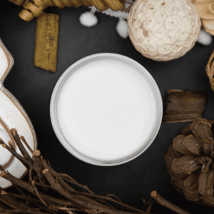 An open jar of white paint sitting on a black table with a brown pine cone, brown sticks, white flowers and brown bark positioned around the jar. 