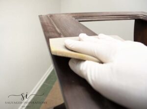 A picture of a hand in a white rubber glove sanding the top of a brown table with a white sanding sponge.