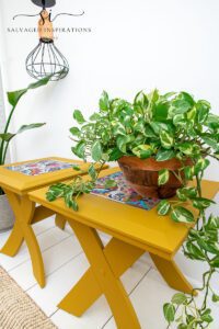 Two yellow rectangular side tables sitting on a white wooden floor with a tan rug in front. The center of the top of the tables has a colorful floral image, a light hangs down with a chain, a green plant in a bronze colored pot sits on top of one table, and a green plant in a white pot sits on the floor to the left of the tables.