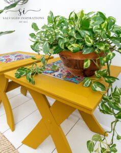 Two yellow rectangular side tables sitting on a white wooden floor with a tan rug in front. The center of the top of the tables has a colorful floral image, a light hangs down with a chain, a green plant in a bronze colored pot sits on top of one table, and a green plant in a white pot sits on the floor to the left of the tables.