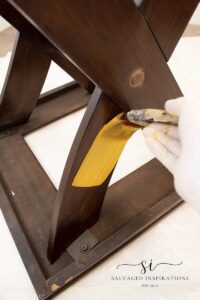 A photo of a brown wooden table sitting on it's top. A hand in a white glove holds a paint brush with yellow paint, the paint brush is painting the table with a yellow swipe.