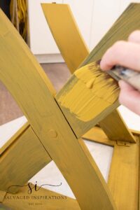 A yellow table sitting upside down on a piece of white paper. A hand is holding a paint brush that is covered with yellow paint, painting one leg of the table.