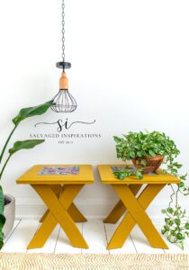 Two yellow rectangular side tables sitting on a white wooden floor with a tan rug in front. The center of the top of the tables has a colorful floral image, a light hangs down with a chain, a green plant in a bronze colored pot sits on top of one table, and a green plant in a white pot sits on the floor to the left of the tables.