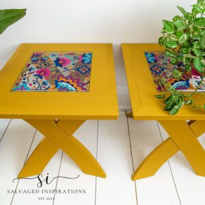 Two yellow rectangular side tables sitting on a white wooden floor with a tan rug in front. The center of the top of the tables has a colorful floral image, a light hangs down with a chain, a green plant in a bronze colored pot sits on top of one table, and a green plant in a white pot sits on the floor to the left of the tables.