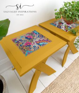 Two yellow rectangular side tables sitting on a white wooden floor with a tan rug in front. The center of the top of the tables has a colorful floral image, a light hangs down with a chain, a green plant in a bronze colored pot sits on top of one table, and a green plant in a white pot sits on the floor to the left of the tables.