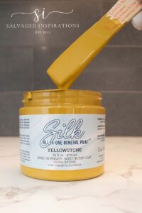 A picture of a yellow jar of paint with a white label that reads Silk All-In-One Mineral Paint Yellowstone, the jar is open and there is a wooden stick above the jar that is covered with yellow paint.