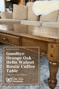 A brown wooden coffee table sitting on a blue and gray rug. A green plant sits on top of the table in a black pot. A tan sofa with a white blanket draped over it is behind the table. In front of the table is a brown box with the words Guest Blog, Goodbye Orange Oak - Hello Walnut Rustic Coffee Table Creative Moments printed in white.