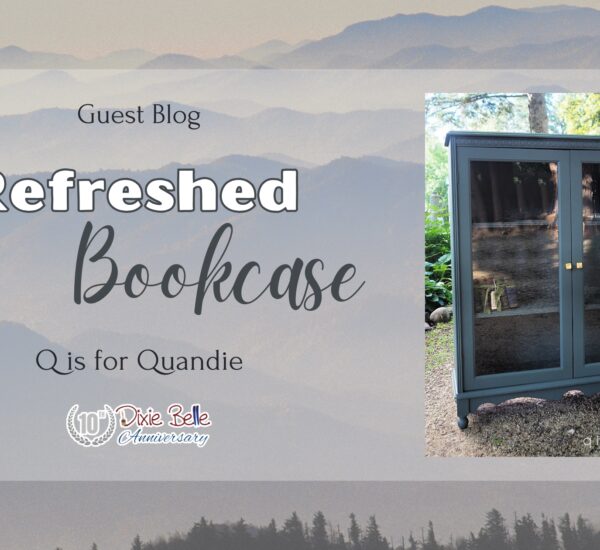 A tall gray painted cabinet with glass doors that have gold knobs sitting on a dirt ground with trees and greenery in the background is to the right of the words Guest Blog Refreshed Bookcase Q is for Quandie. The background is the Great Smoky Mountains.