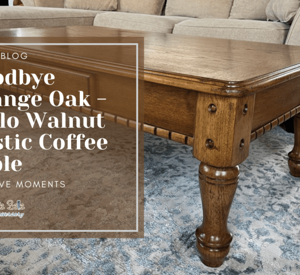 A brown wooden coffee table sitting on a blue and gray rug. A green plant sits on top of the table in a black pot. A tan sofa with a white blanket draped over it is behind the table. In front of the table is a brown box with the words Guest Blog, Goodbye Orange Oak - Hello Walnut Rustic Coffee Table Creative Moments printed in white.