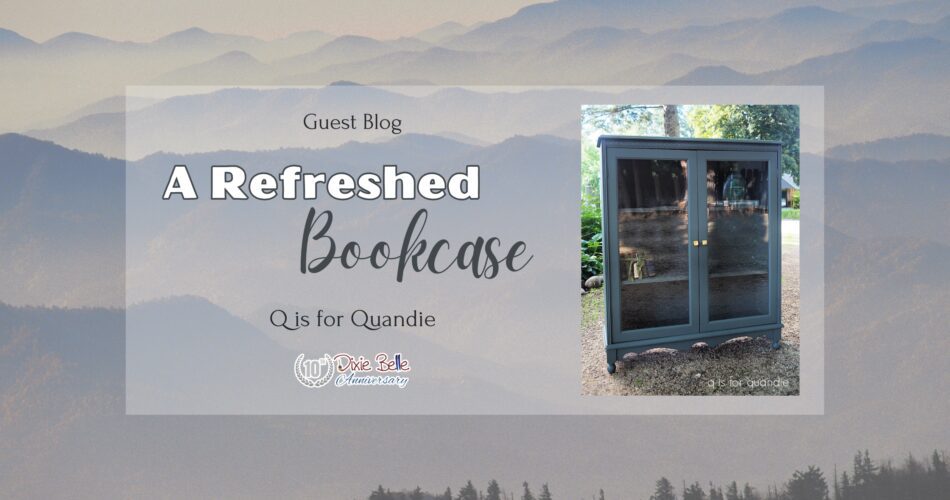 A tall gray painted cabinet with glass doors that have gold knobs sitting on a dirt ground with trees and greenery in the background is to the right of the words Guest Blog Refreshed Bookcase Q is for Quandie. The background is the Great Smoky Mountains.