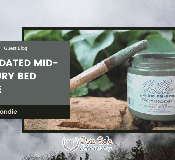 An open jar of Smoky Mountains Silk All-In-One Mineral Paint sitting on a brown rock with greenery in the background. A paint brush with paint on the bristles sits across the top of the jar. The words "an updated mid-century bed frame" are printed to the left of the paint jar. The entire background of the picture is a photo of the Great Smoky Mountains.