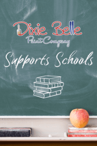 A green chalk board with Dixie Belle Paint Company logo, Supports Schools and a stack of 4 books on the chalk board. The tray below the chalkboard holds a black eraser, a red eraser and a red apple.