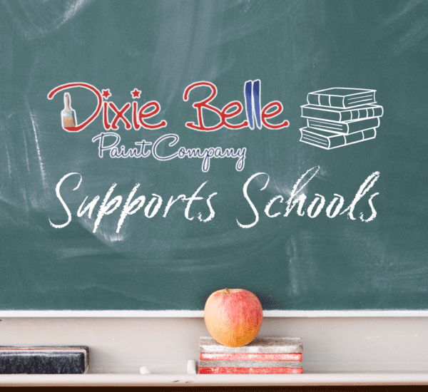 A green chalk board with Dixie Belle Paint Company logo, Supports Schools and a stack of 4 books on the chalk board. The tray below the chalkboard holds a black eraser, a red eraser and a red apple.