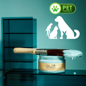 An open jar of Dixie Belle Blue paint sitting on a blue table with a paint brush that has paint on it lying across the top of the jar. Above the jar is a paw print in green with the words Pet Friendly to the right of the paw print and a white shadow of a bird, cat, dog and bunny sitting underneath the paw print.