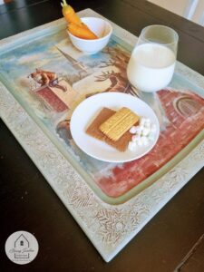 a white painted tray with Santa on the Rooftop decoupage paper applied to the top of it. Sitting on the tray are a white bowl with two orange carrots, a glass with white milk, and a white plate with chocolate, a graham cracker and marshmallows on it.