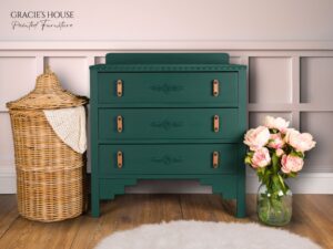 a dresser painted with Midnight Green Silk All-In-One Mineral Paint sits on a brown wooden floor with a white furry rug in front of it. A brown wicker basket is on the left side of the dresser; a clear vase with roses in it sits on the floor to the right of the dresser.