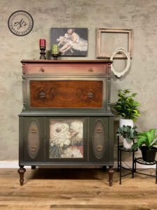 A photo of a dresser painted green and pink with a brown wooden drawer staged on a brown wooden floor in front of a brown marblish wall with green plants to the right of it.