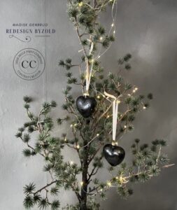 Two heart shaped ornaments painted black with gold ribbons hanging from a green tree in front of a gray wall