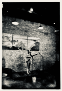 Meghan D standing on a stage in front of a brick wall singing with her guitar