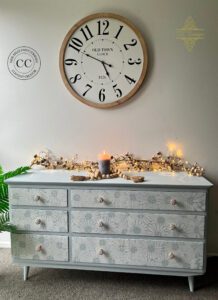 A dresser painted with Harbor Silk All-In-One Mineral Paint and the Crazy Daisies stencil sits on a light brown floor in front of a white painted wall. A large round clock hangs on the wall above the dresser.