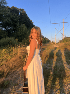 Meghan D wearing a white dress and holding her guitar in her left hand in a field with brush and trees