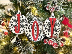 3 Christmas tree ornaments painted white with black snowflakes, together they spell JOY painted red, hanging on a Christmas tree