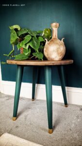 A small table with Midnight Green painted legs sitting on a white floor in front of a green painted wall with white trim; on top of the table is a green plant and a painted vase