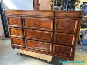 A photo of a brown wooden dresser with the hardware removed, sitting on a wooden cart