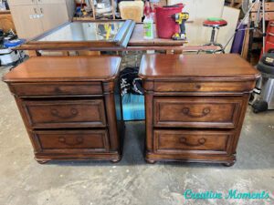 Two brown wooden nightstands with the hardware removed sitting on a gray cement floor. Various tools are in the background.