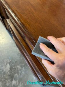 A brown wooden dresser top being sanded by a hand holding a gray sanding sponge.