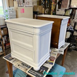 A pair of white painted nightstands sit on top of a table covered with newspapers.