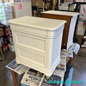 A pair of white painted nightstands sit on top of a table covered with newspapers.