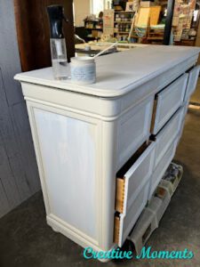  A dresser that has been painted with Bonding BOSS and partially painted with Endless Shore Silk All-In-One Mineral Paint. A spray bottle and open jar of Endless Shore Silk paint sits on top of the dresser.