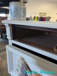 A closeup picture of a white painted dresser drawer partially open with a hand holding a paint brush in front and an open jar of Endless Shore Silk All-In-One Mineral Paint sitting on top of the dresser.