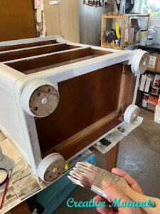 A white painted nightstand that has the drawers removed sits on it's back with a hand holding a paint brush next to the circular feet 