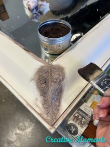 The corner of a white painted mirror frame with Brown Best Dang Wax applied to the ornamental part; an open can of brown Best Dang Wax sits on the mirror and a hand holding a paint brush is over the frame