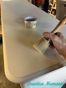 A white painted dresser top with an open can of clear Best Dang Wax and a hand holding the Best Dang Brush on top of the dresser.