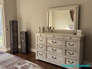 A white painted dresser with a mirror on top sits on a brown wooden floor, in front of an off white painted wall. A flowery rug sits on the floor in front of the dresser. Two rectangular brown pieces of decor sit on the floor to the left of the dresser.