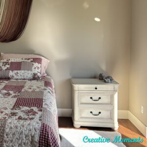 A white painted nightstand sits on a brown wooden floor in front of a tan painted wall; to the left is a bed with a pink and flowery quilted bespread 