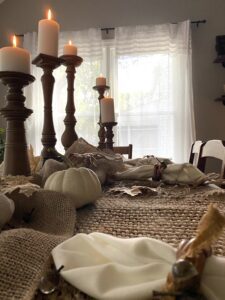 A table with burlap placemats, a white napkin, a white painted pumpkin and brown painted candlesticks holding white candles.