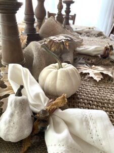 A white napkin tied with a brown piece of material is sitting on a piece of burlap with white painted gourd and pumpkin around it, brown leaves and brown painted candlesticks are in the background