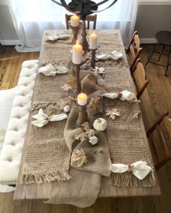 A brown wooden table with burlap place mats, white painted pumpkins and gourds, white napkins, brown stained candlesticks holding white candles.