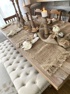 A brown wooden table with burlap place mats, white painted pumpkins and gourds, white napkins, brown stained candlesticks holding white candles.