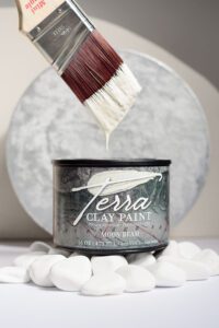 A container of Moon Beam Terra clay paint sitting on white rocks in front of a white painted circle with a paint brush above the jar that has paint on the bristles.