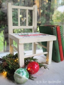 A white painted wooden chair sitting on a gray floor with green an blue books leaning on the right side, green and white Christmas balls and green lit holly are on the floor to the left of the chair.