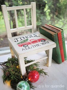 A white painted wooden chair sitting on a gray floor with green an blue books leaning on the right side, green and white Christmas balls and green lit holly are on the floor to the left of the chair. On the seat of the chair is a red painted truck with a green painted tree in the back of the truck with Jingle All the Way painted in black