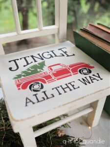 Top view of a white painted wooden chair that has a red painted truck with a green painted tree in the back of the truck; Jingle All The Way is painted on the chair in black