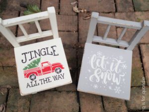 Top view of 2 painted wooden chairs. The left chair is painted white with a red painted truck with a green painted tree in the back of the truck, Jingle All The Way is painted with black; the right chair is painted tan with the words let it snow and snowflakes painted in white on the seat. Both chairs are sitting on a brown brick ground.