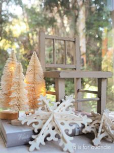 A tan painted wooden chair with 3 off-white trees to the left of the chair and white painted wooden snowflake in front of it.